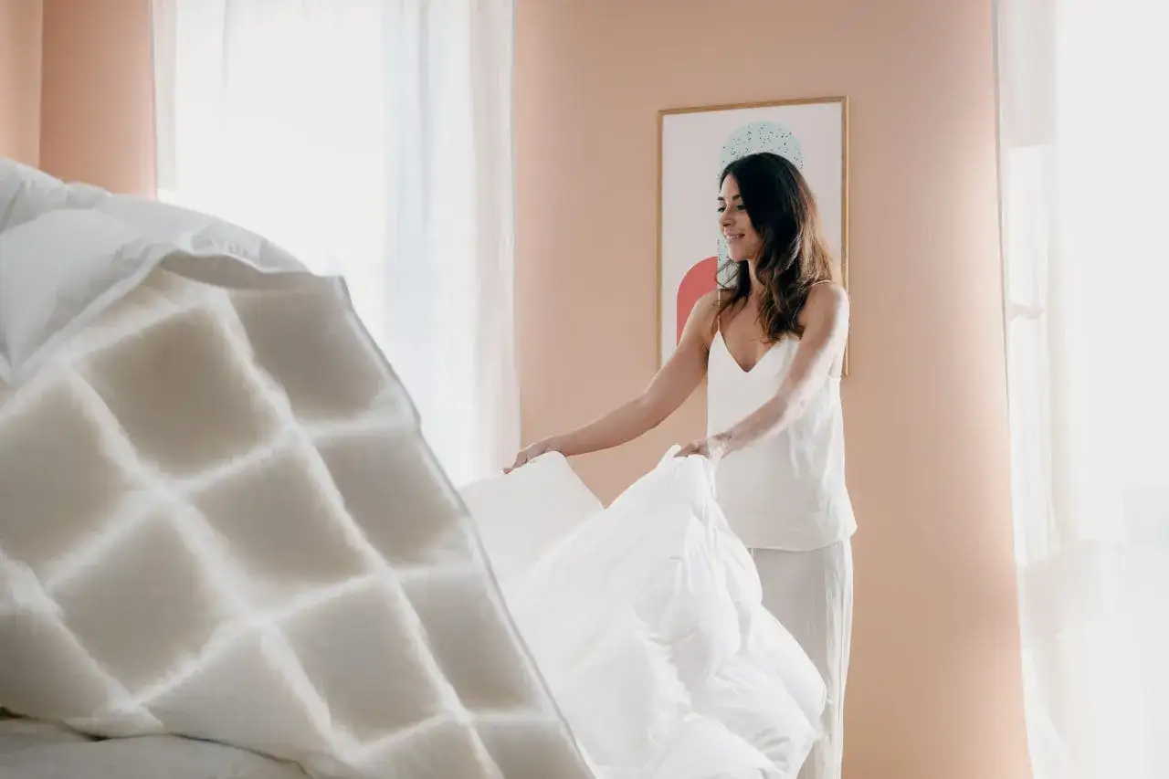 Donna sorride mentre sistema un soffice piumone bianco, mostrando come piegare un piumone per un letto ordinato.