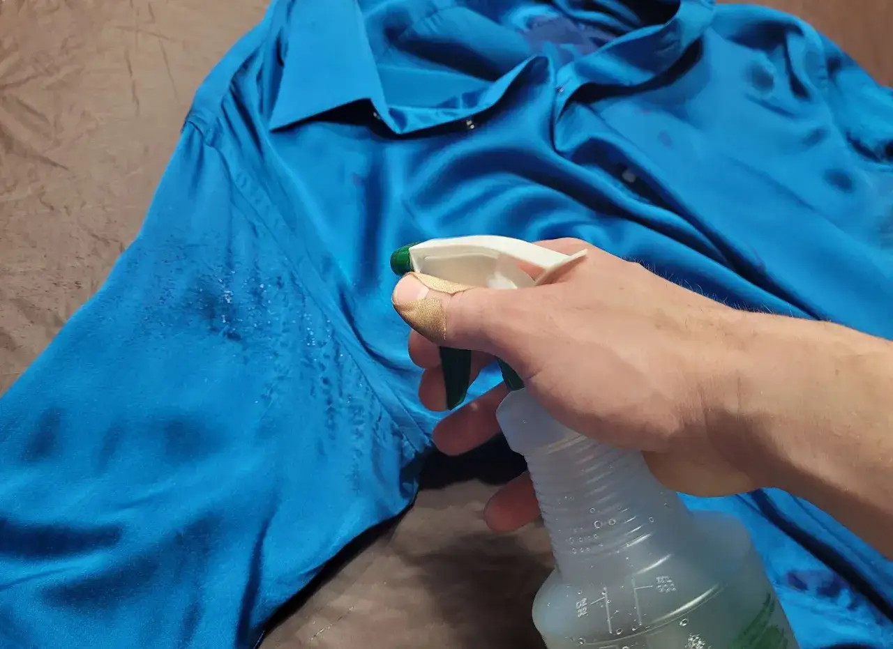 Mano con cerotto spruzza un prodotto su una camicia di seta blu per togliere macchie di sudore vecchie.