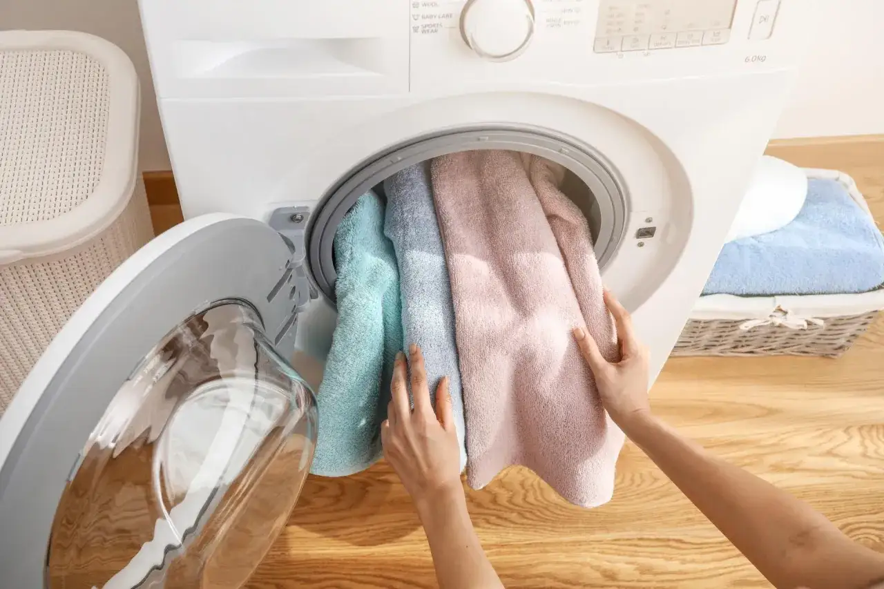 Mani che inseriscono asciugamani color pastello in una lavatrice, un passo fondamentale per imparare come lavare asciugamani in lavatrice.