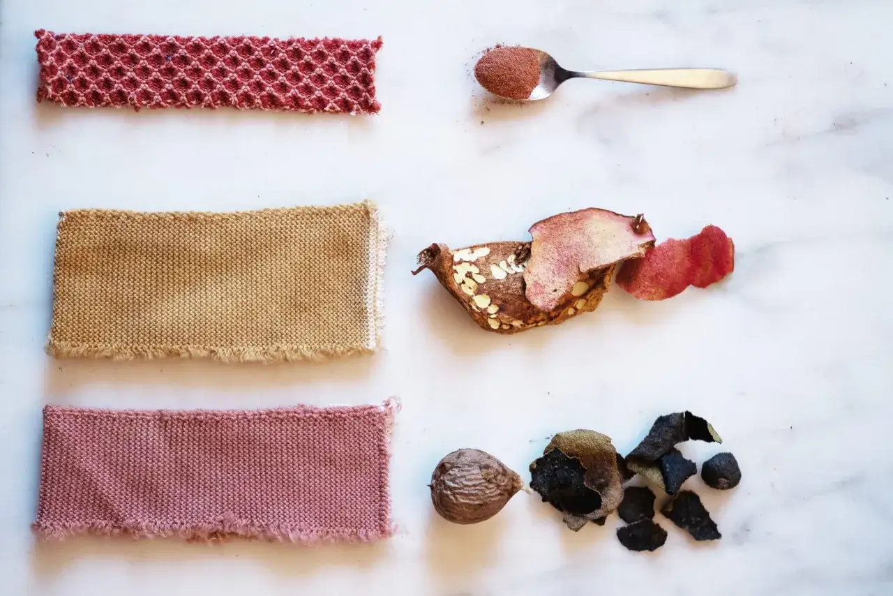 Campioni di tessuto tinti con colorante naturale per tessuti: rosa, beige e rosso. Accanto, ingredienti naturali come melograno, noce e scorze.