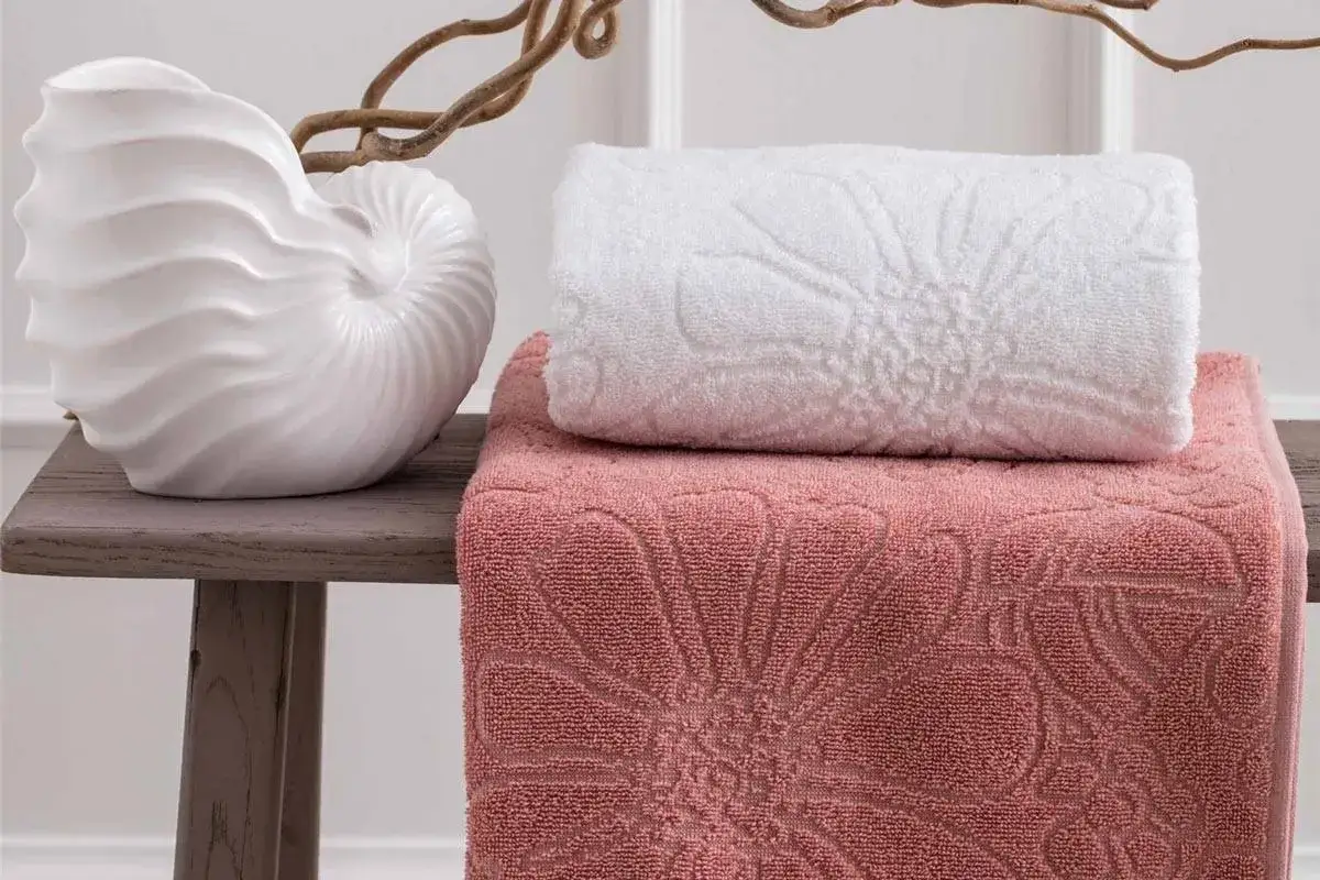 Asciugamani rosa e bianchi con motivi floreali in rilievo, disposti su un tavolino con una decorazione a conchiglia. La grammatura degli asciugamani ne garantisce morbidezza.