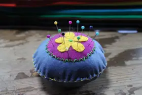Puntaspilli fai da te a forma di fiore, con petali gialli su sfondo viola e base blu, decorato con cuciture a zig-zag verdi e gialle.