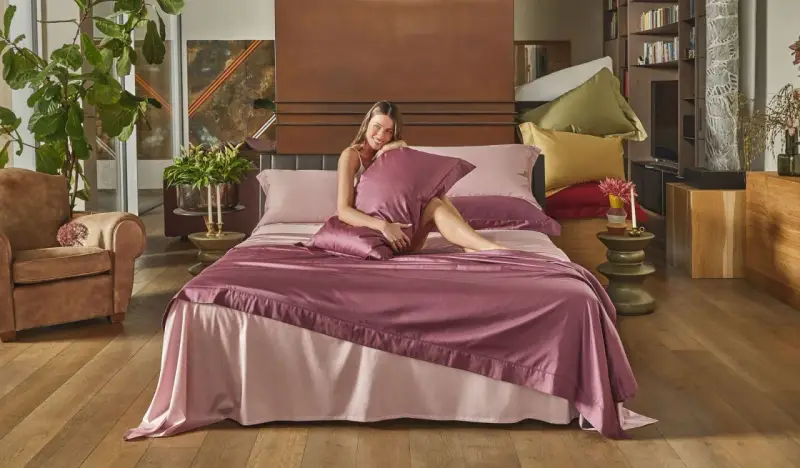 Donna sorridente su un letto con lenzuola rosa e copriletto prugna, che illustra la differenza tra piumone e trapunta.