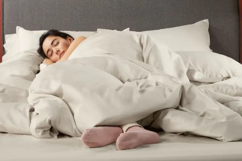 Donna dorme avvolta in lenzuola in microfibra che sembrano farla sudare, con solo i piedi in calzini rosa visibili.