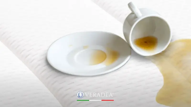 Tazza rovesciata su un materasso bianco, con liquido marrone versato. Scopri come disinfettare il materasso per eliminare macchie e odori.
